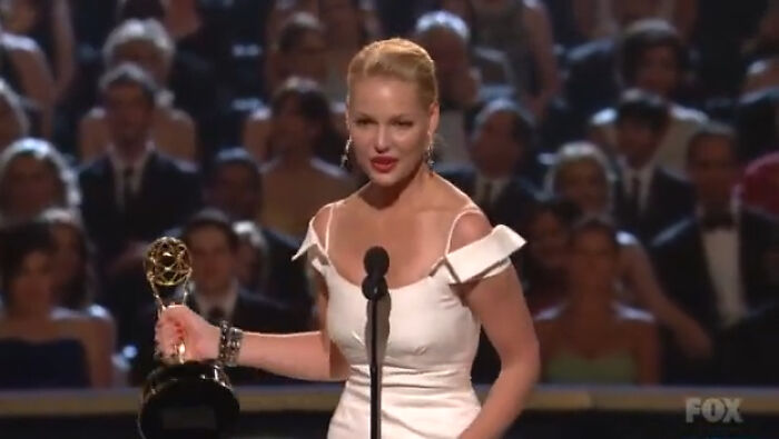 Actor in a white dress accepting an award on stage during a scandalous awards show moment.
