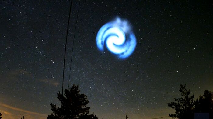 Mysterious spiral light in European night sky, seen amid a starry background with silhouetted trees below.