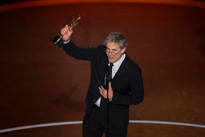 Man in a suit holding an Oscar, speaking at the Oscars 2025 ceremony stage.