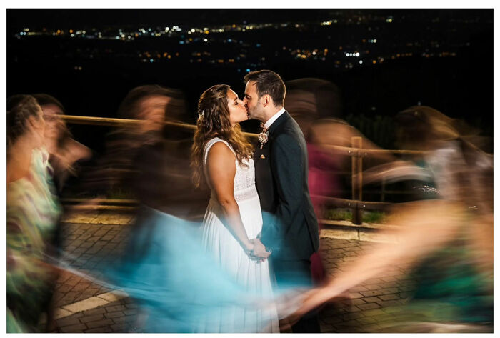A couple shares a romantic kiss at night with blurred guests dancing around, captured in an outstanding wedding photograph.