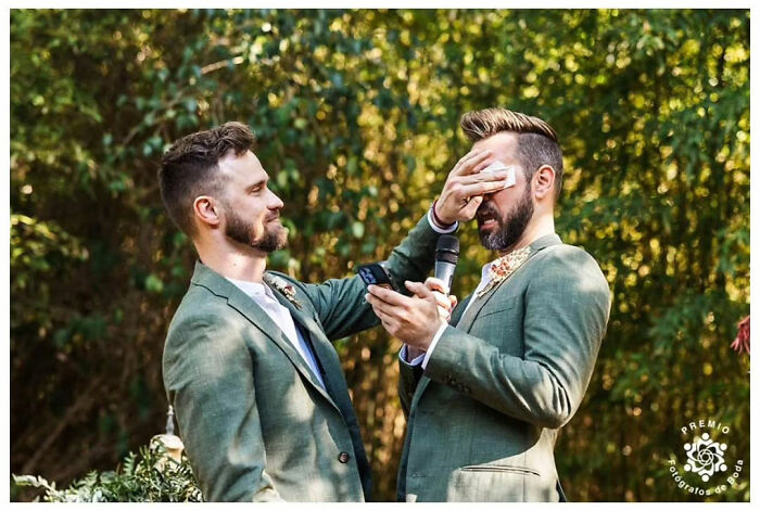 Two grooms in green suits share an emotional moment during their wedding ceremony, one wiping tears with a tissue.