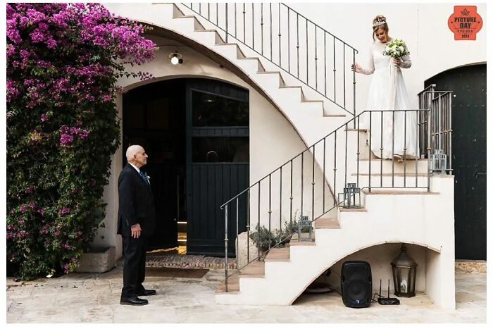 Bride descends stairs in wedding dress, holding bouquet, while elderly man watches; a captured moment of love by Jordi Tudela.