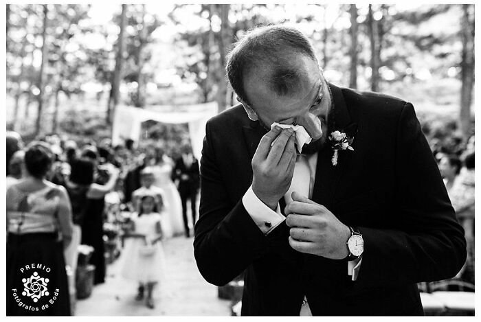 Groom wiping tears during a wedding ceremony, surrounded by guests and a forest backdrop, capturing a precious moment of love.