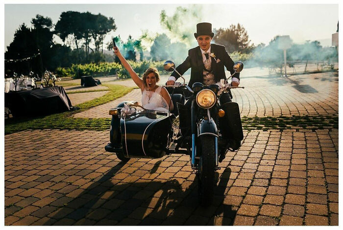 Bride and groom on a vintage motorcycle with sidecar, celebrating a joyful wedding moment outdoors.
