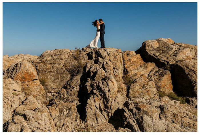 A couple embraces on rocky cliffs under a clear blue sky, capturing a precious wedding moment of love.