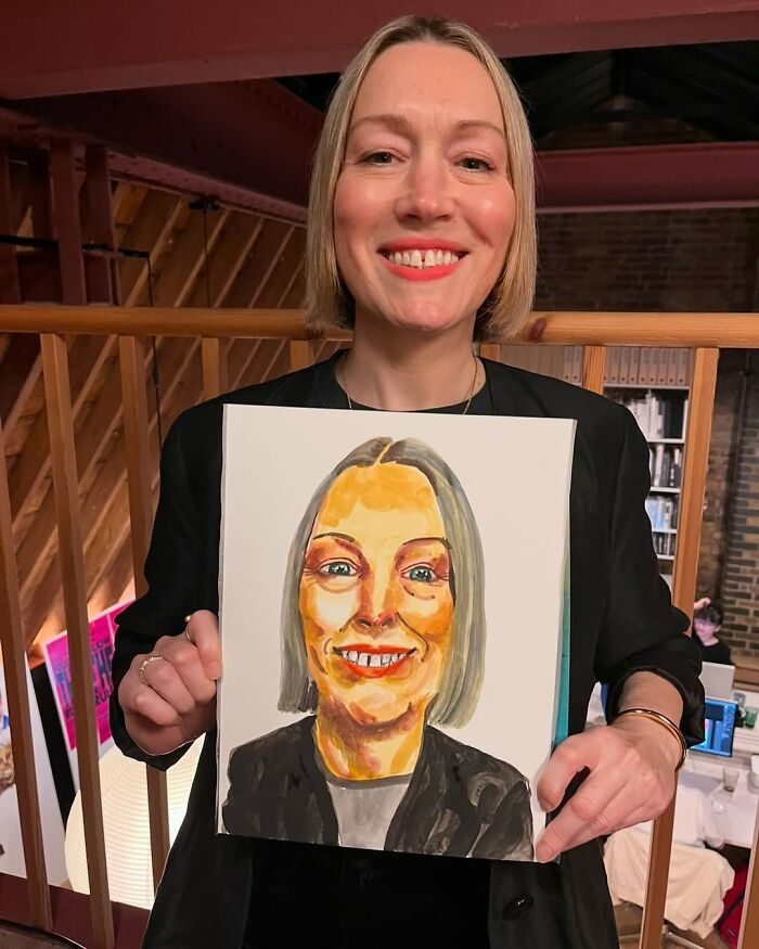 Smiling woman holding an expressive portrait by a British artist in a rustic interior setting.