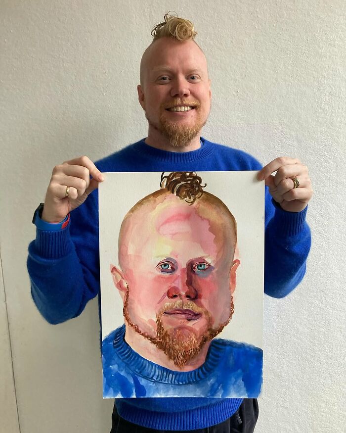 British artist showcasing an expressive portrait he painted, wearing a bright blue sweater and smiling.
