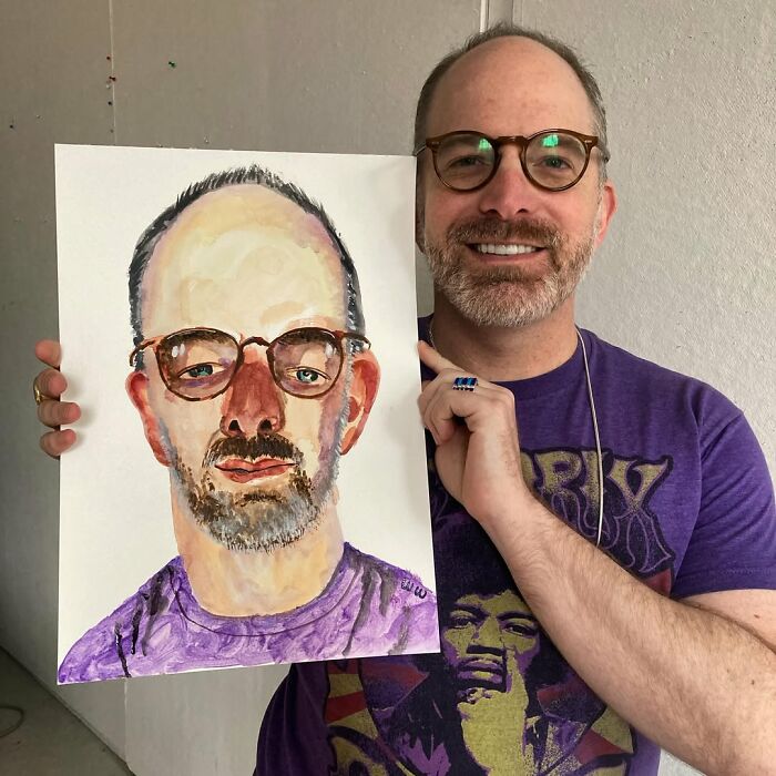 British artist holding an expressive portrait of himself, wearing glasses and a purple shirt.
