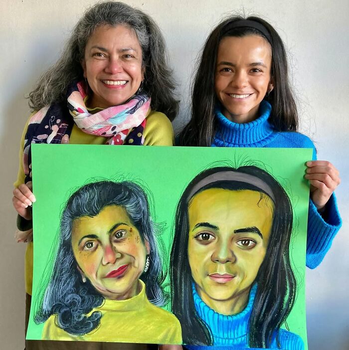 Two women smiling, holding expressive portraits by British artist against a white wall.