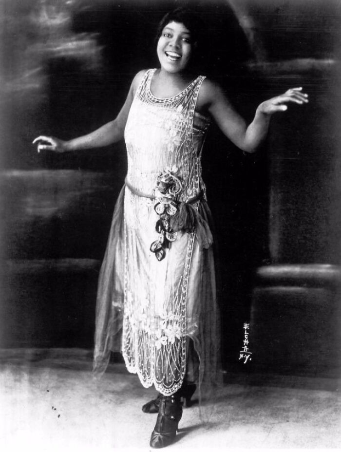 A woman in a vintage dress, showcasing 1920s fashion, smiles with arms slightly raised, highlighting how much the world has changed.