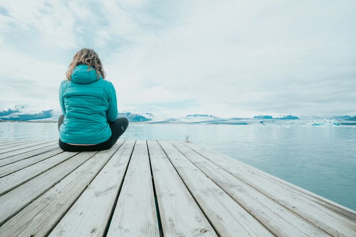 Person in a turquoise jacket sitting on a wooden dock, gazing at a vast serene lake, reflecting on lost friendship.