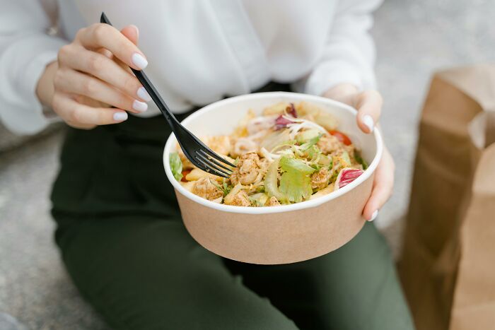 Person holding a salad bowl, using a black fork to eat outdoors, highlighting healthy food traits.