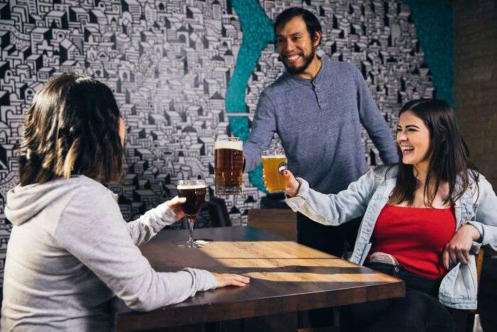 Three friends enjoying drinks, sitting at a wooden table, smiling in a lively bar setting with a mural background.