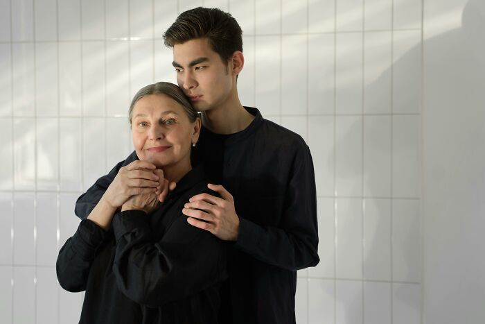 A young man and older woman, both in black outfits, stand closely in a tiled room, representing varied human traits.