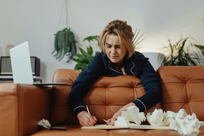 A woman in pajamas on a couch, writing with crumpled papers nearby.