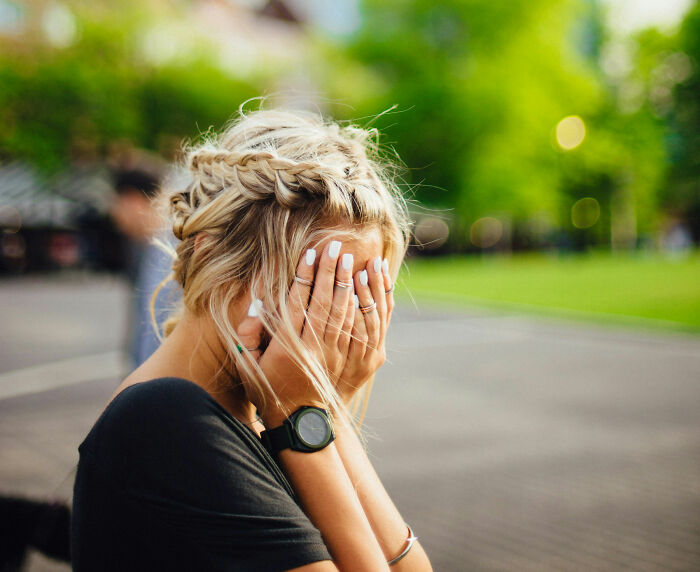 A woman covers her face with her hands, standing in a park, looking scared.