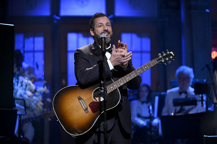 Adam Sandler performing with a guitar on stage, highlighting comedy's lucrative impact.