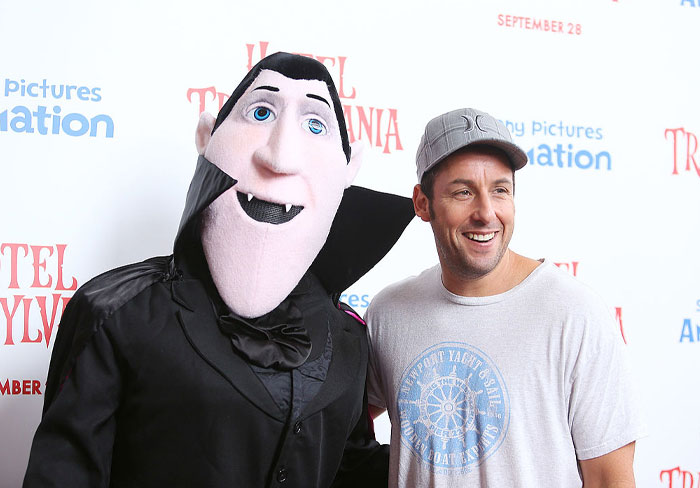 Adam Sandler smiles next to a Hotel Transylvania character at a movie event, showcasing comedy's financial success.