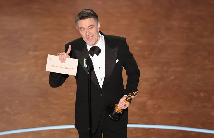 Man in tuxedo holding Oscar trophy on stage at the 2025 Oscars ceremony.
