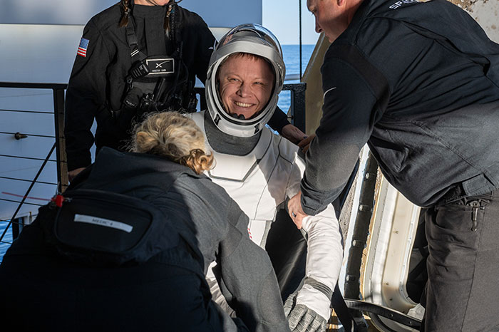 Astronaut being assisted by doctors, smiling in a space suit, with concern for walking ability.