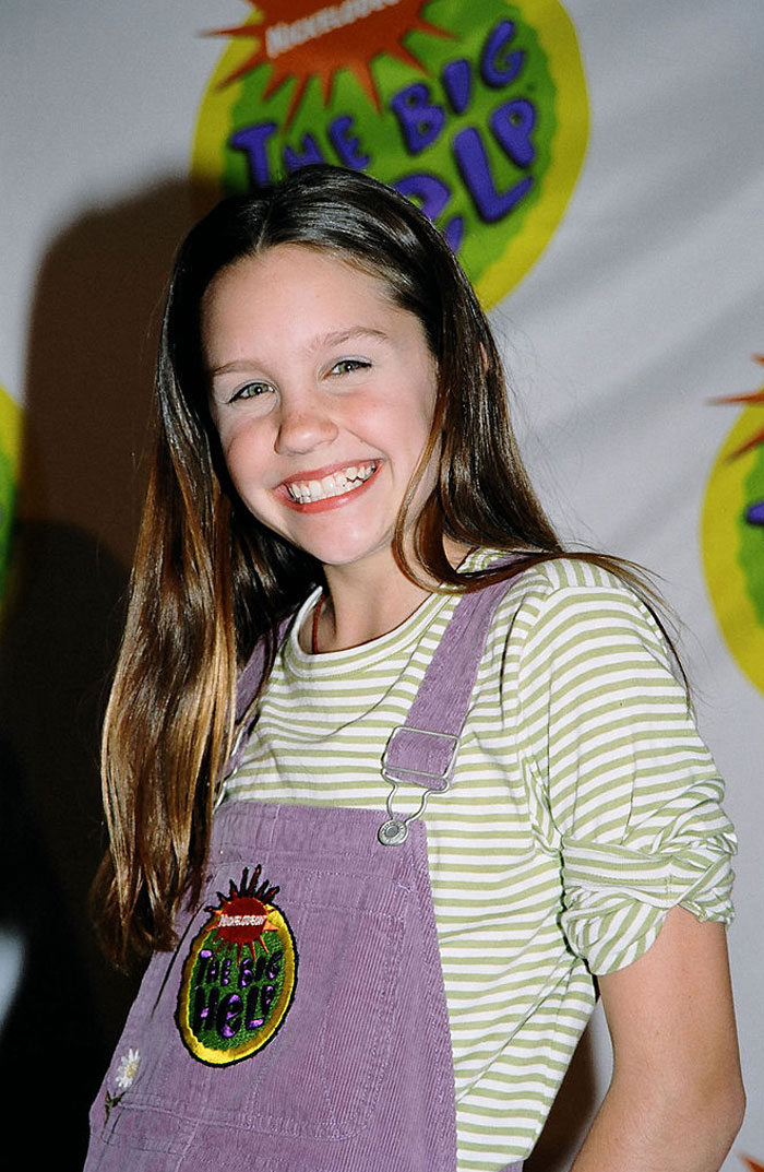 Smiling young Amanda Bynes in striped shirt and purple overalls, posing at an event backdrop.