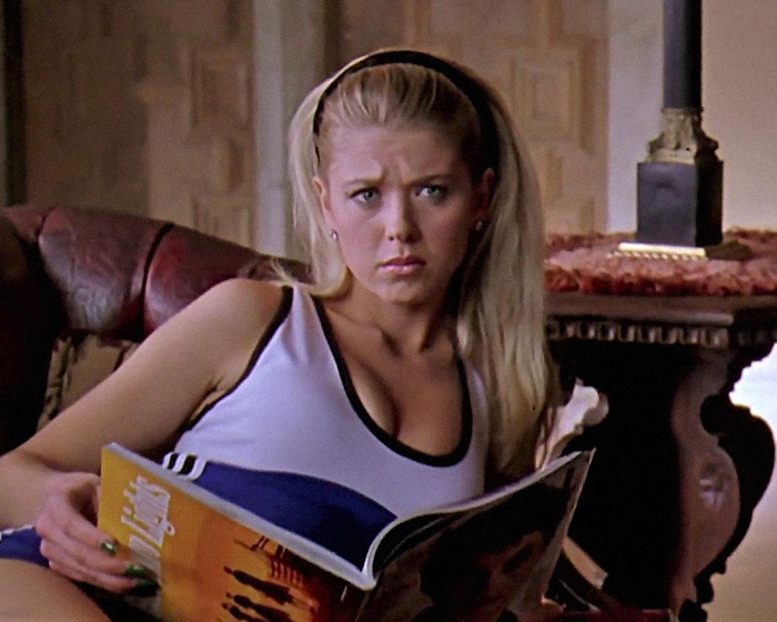 Blonde woman reading a magazine, wearing a white tank top, sitting on a sofa, scene from a movie.