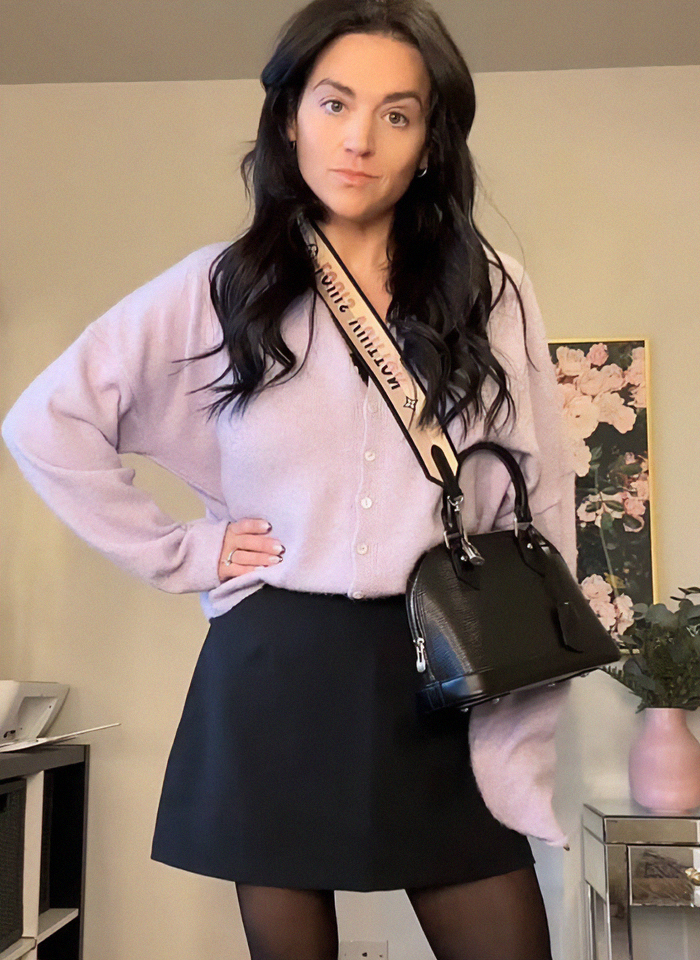 Woman shows dramatic Ozempic transformation with excess skin, wearing lilac sweater and holding a handbag indoors.