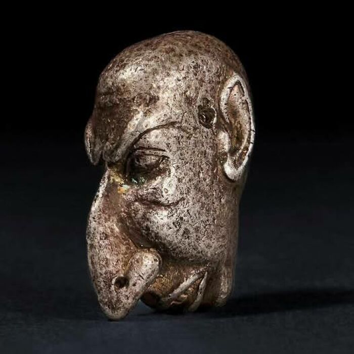 Ancient history artifact, a carved metal figurine with a unique face shape, set against a dark background.