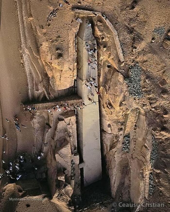 Ancient history site with people exploring a large, partially carved stone obelisk in a sandy area.
