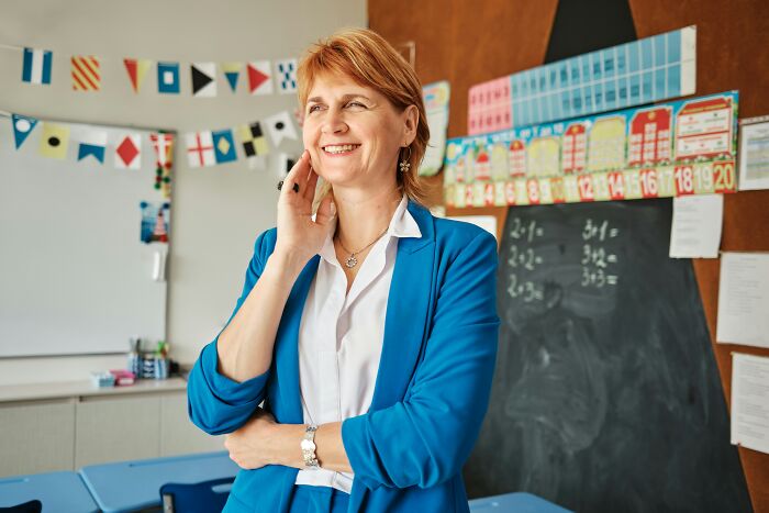 Smiling teacher in a blue blazer standing in a colorful classroom, embodying things that receive undue hate.