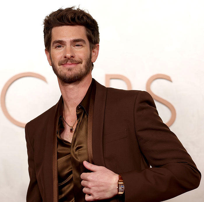 Man in a brown suit at the Oscars, smiling, with short hair and a brown shirt; Oscars event backdrop visible.