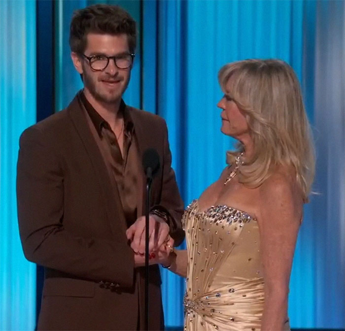 Actor in a brown suit holding hands with a woman in a gold dress on stage, engaging in a heartfelt moment.