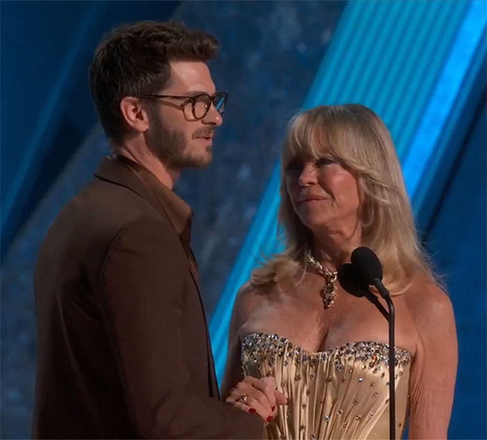 Man in glasses with Goldie Hawn, sharing an emotional moment on stage at an event.
