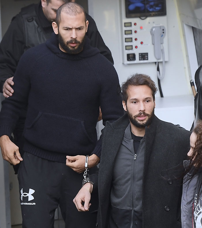 Two men, one in a black sweater and one in a black coat, walking while handcuffed together.