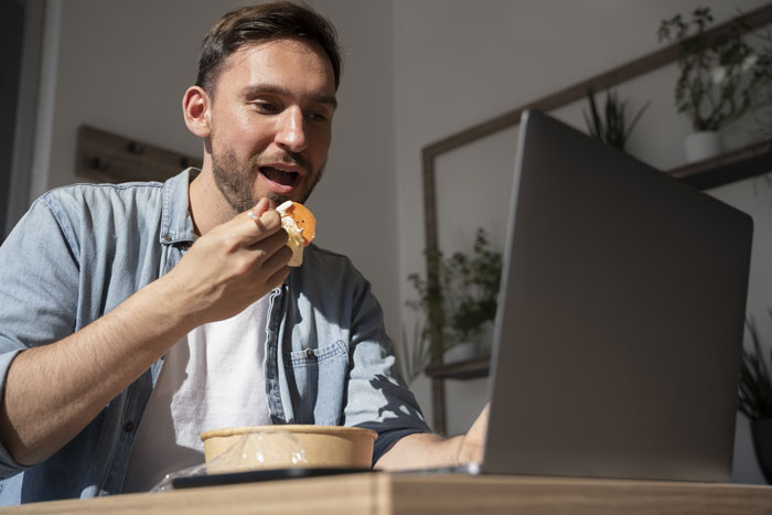 Man in breakroom eating during work, sitting by a laptop, casually dressed.