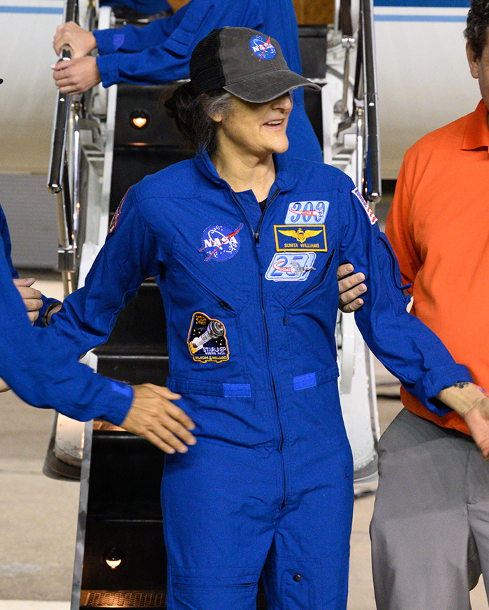 NASA astronaut in blue suit and cap, being assisted while exiting a spacecraft.