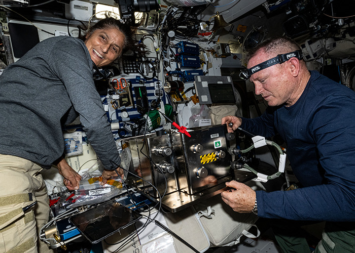 Astronauts working on the International Space Station, handling equipment and smiling amidst complex machinery.