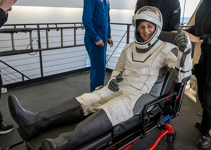 Astronaut in spacesuit giving thumbs up, seated on stretcher post-landing, with medical personnel nearby.