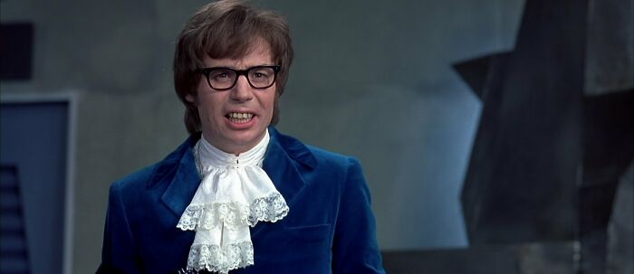 Actor in blue velvet suit and frilly shirt speaks passionately in a dimly lit, angular room.