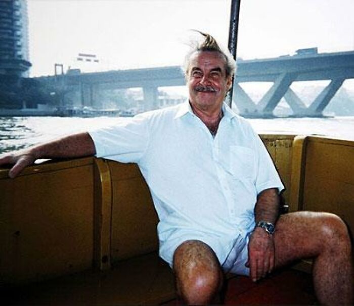 Man smiling on a boat with a bridge in the background, wearing a white shirt; part of creepy photos collection.
