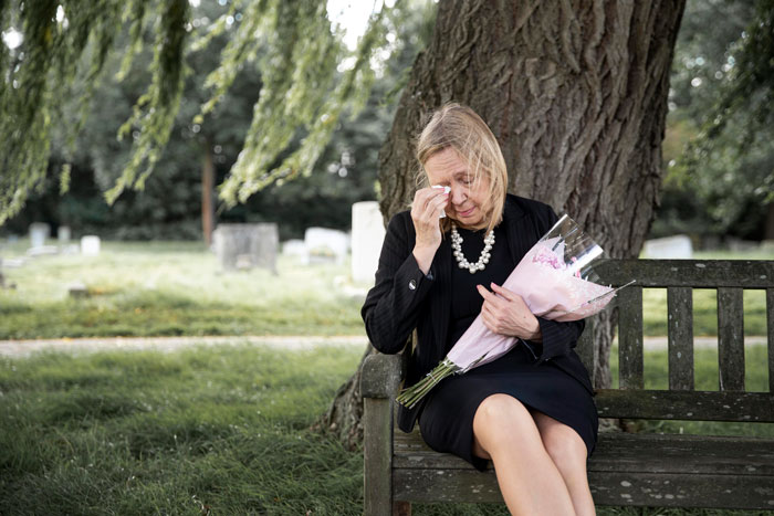 Woman sitting on a bench in a cemetery, holding flowers, wiping tears, reflecting on naming baby after her mother.