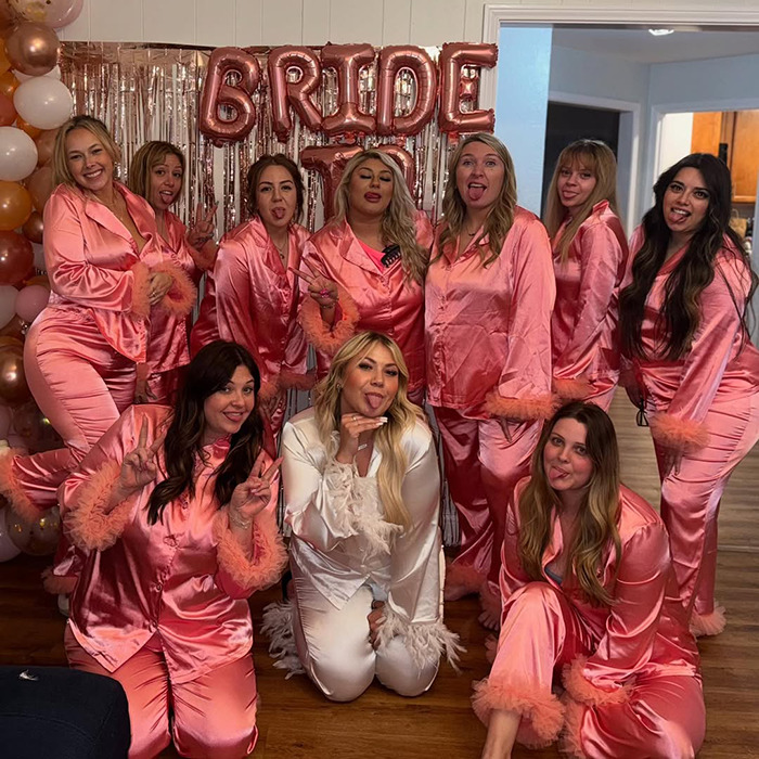 Bride-to-be in white poses with friends in pink pajamas at her bachelorette party, under "bride" balloons.