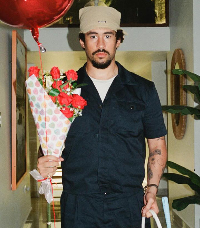 Man in dark outfit holding roses and a balloon indoors, related to Bad Bunny Calvin Klein ad campaign.
