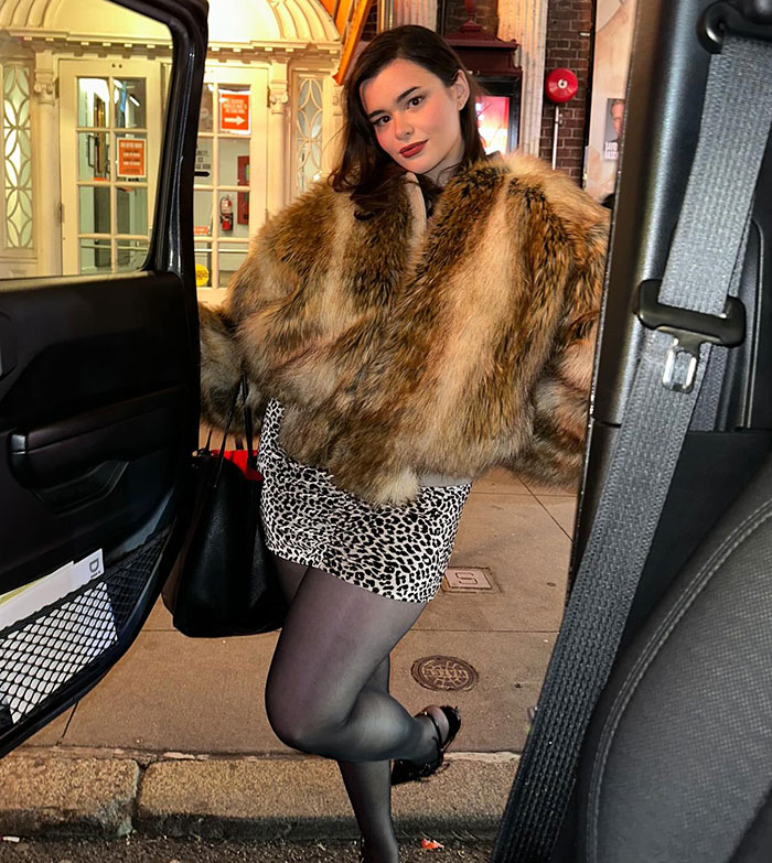 Barbie Ferreira in a fur coat and leopard skirt, standing by a car on a city street.