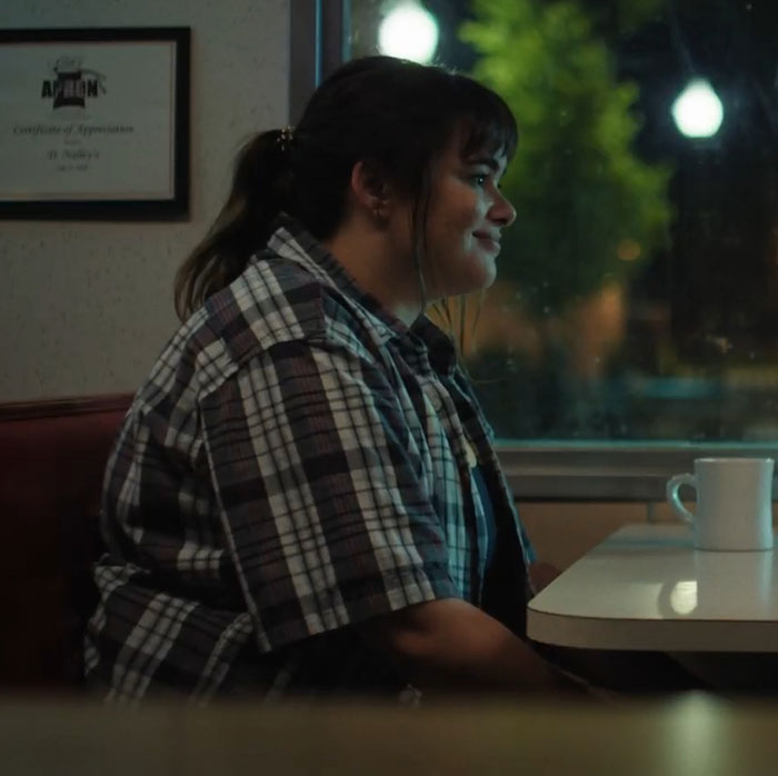 Barbie Ferreira in a scene filmed pre-weight loss, sitting in a diner, wearing a plaid shirt, and smiling.