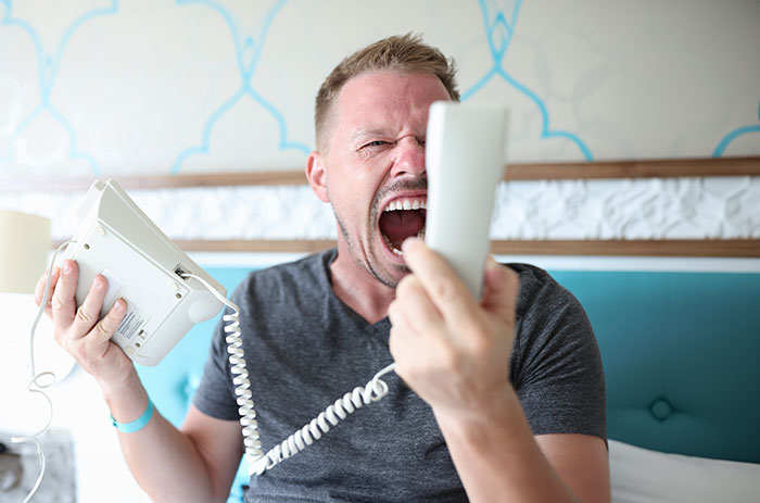 Man in gray shirt shouting into landline phone, demonstrating basic knowledge of phone etiquette.
