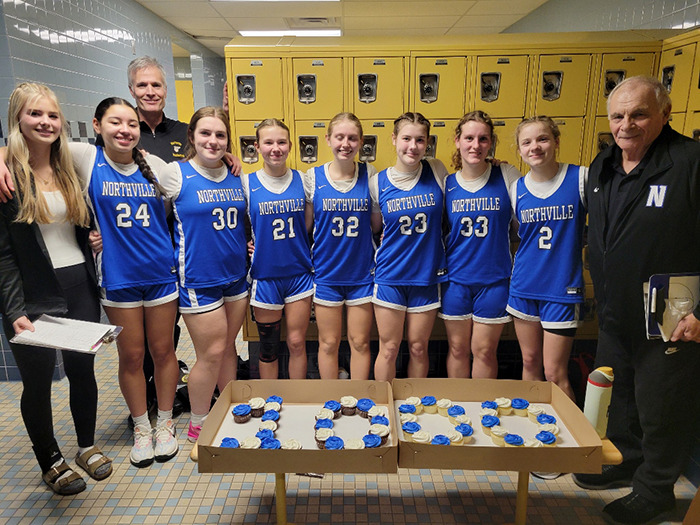Northville basketball team in blue uniforms with coaches in locker room setting, celebrating with cupcakes on the table.