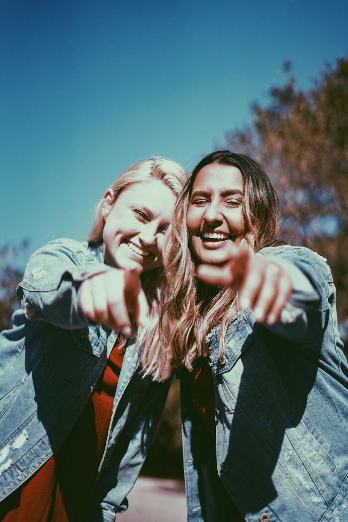 Two smiling women in denim jackets pointing, outdoors on a sunny day.