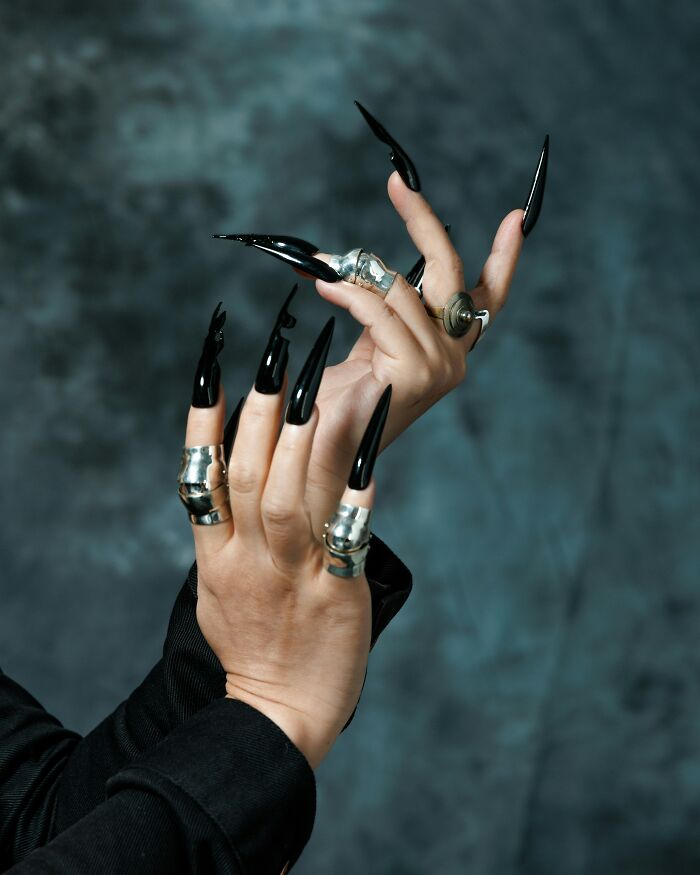 Hands with long black pointed nails and silver rings, showcasing extreme beauty trends.