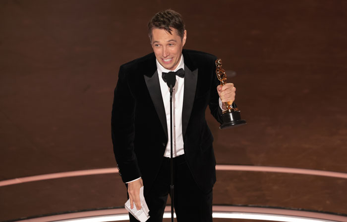 Winner at Oscars 2025 holding an award in a black tuxedo, smiling on stage.